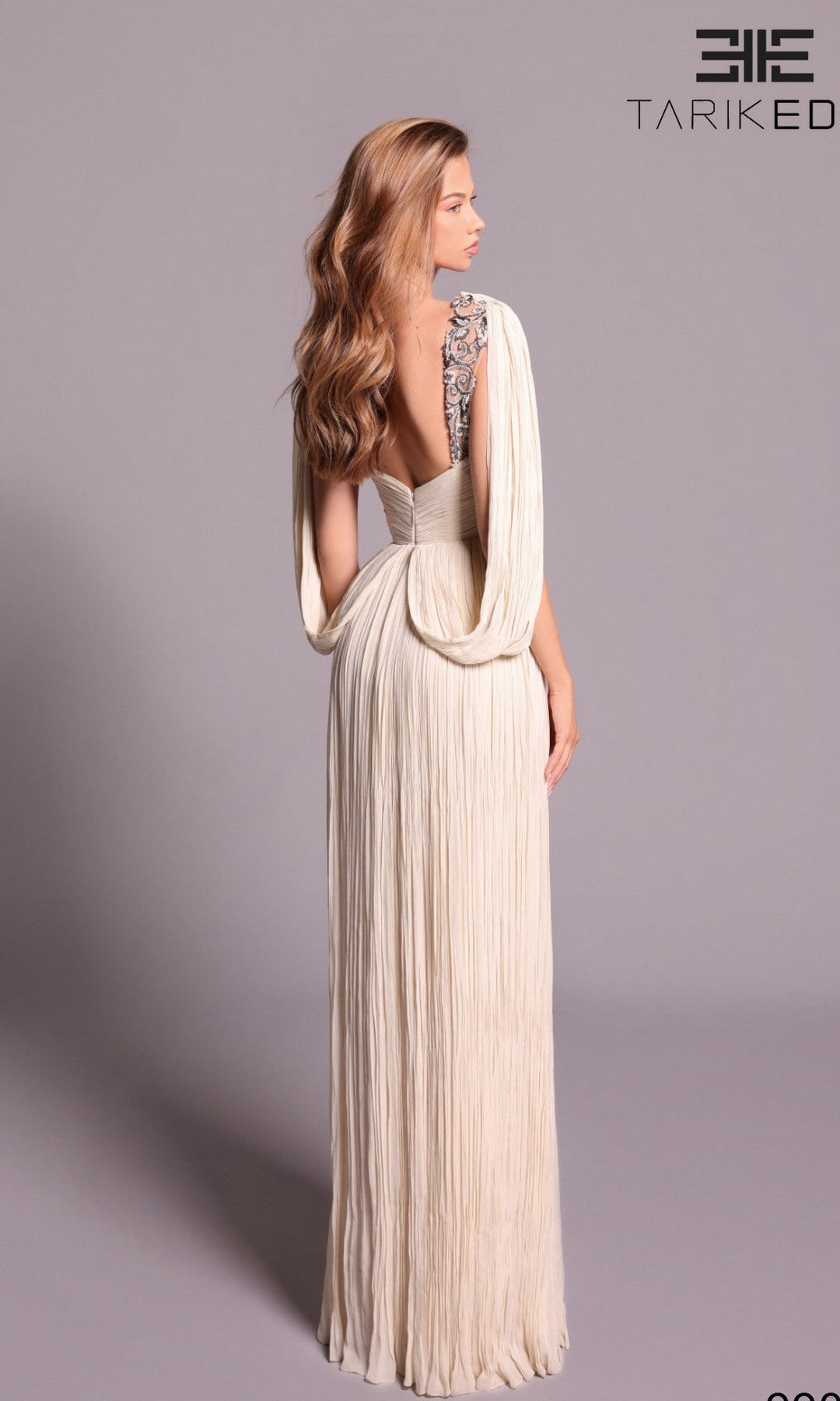 A woman wears the ivory, floor-length Tarik Ediz 99031 gown featuring pleated crepe, draped sleeves, and an embellished shoulder, standing against a plain background with the Tarik Ediz logo in the top right corner.